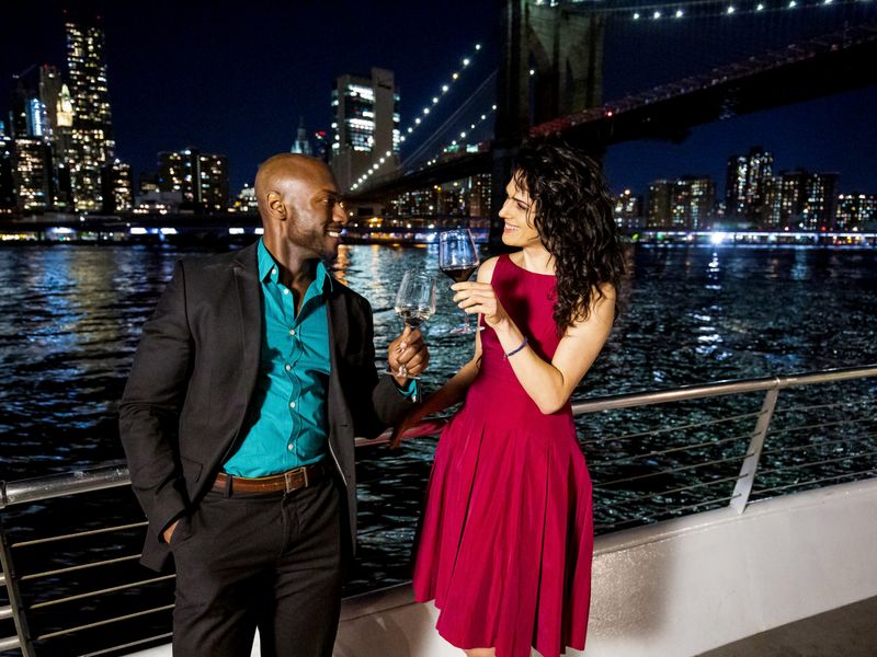 Paar stößt mit Wein auf dem Außendeck eines Schiffes an, während die beleuchtete Brooklyn Bridge und die Skyline von Manhattan im Hintergrund zu sehen sind.