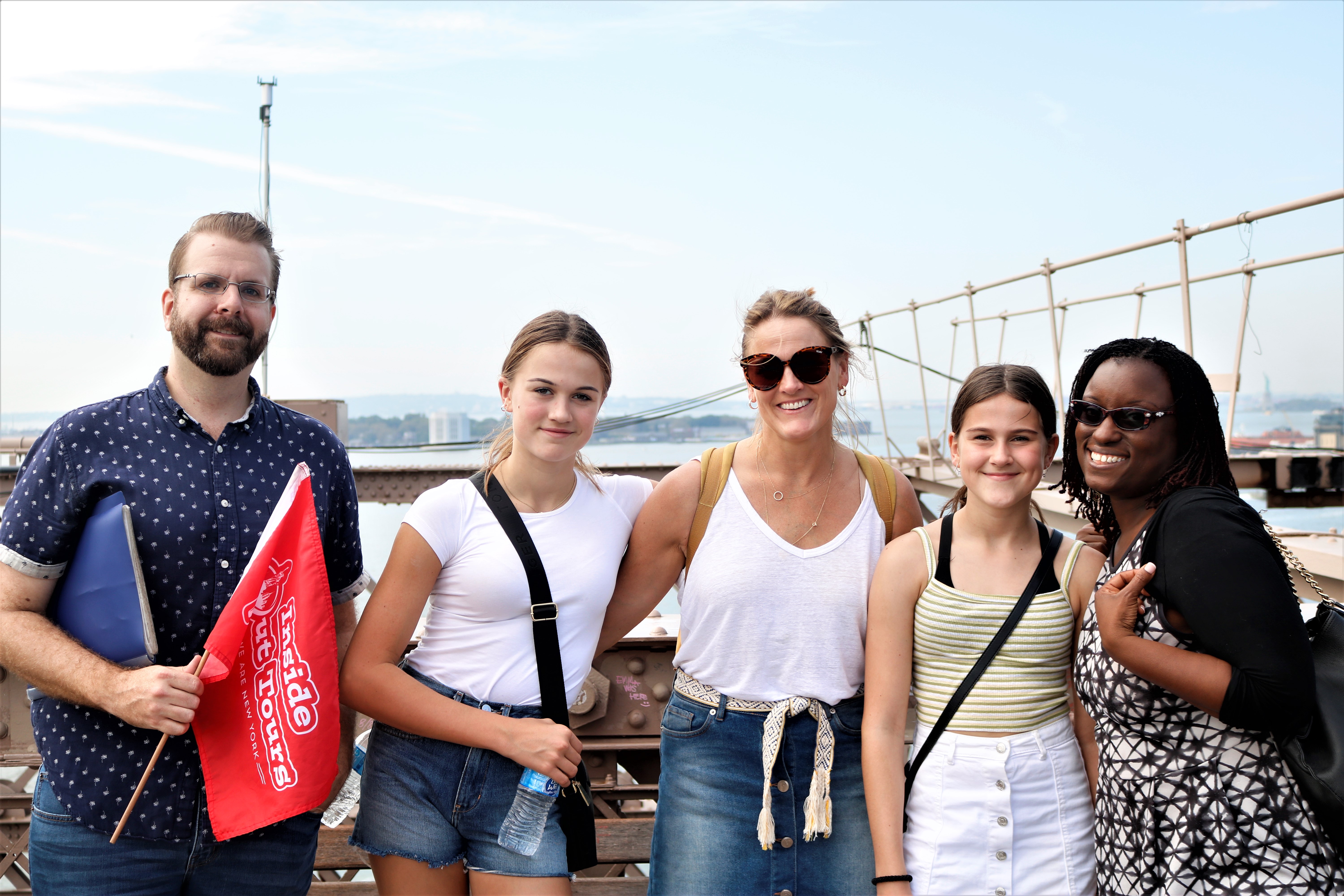 Eine Gruppe von Touristen posiert glücklich für ein Foto auf der Brooklyn Bridge, während ein Teilnehmer eine rote Touristenflagge hält.