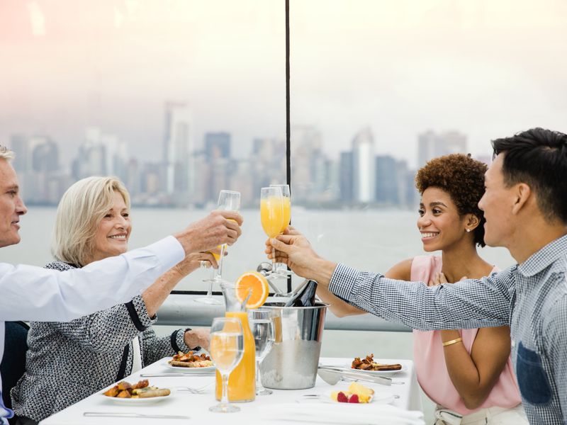 Eine diverse Gruppe von vier Personen stößt fröhlich mit Sektgläsern und Orangensaft bei einem Frühstück an Bord an.