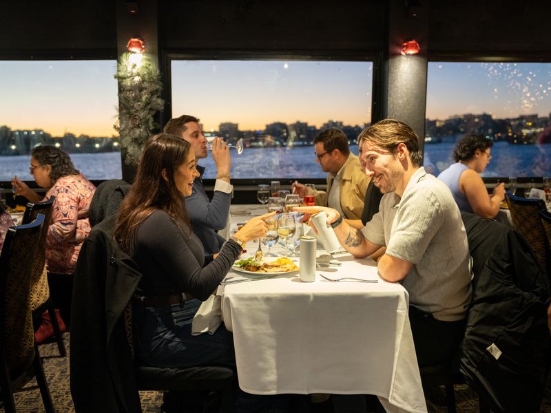 Gäste genießen ein Dinner am gedeckten Tisch mit Blick auf das Wasser und die Skyline bei Sonnenuntergang.