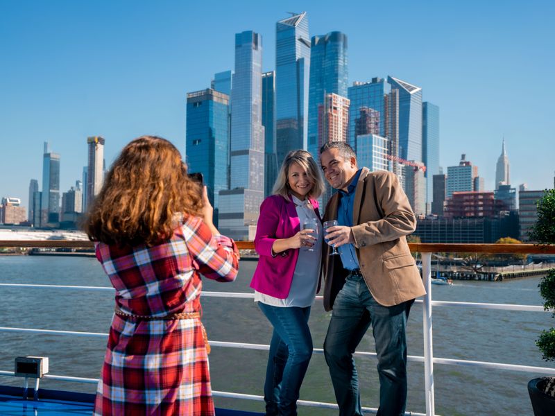 Eine Frau in kariertem Hemd fotografiert ein Paar, das mit Gläsern in der Hand vor der Kulisse einer modernen Wolkenkratzer-Skyline posiert.