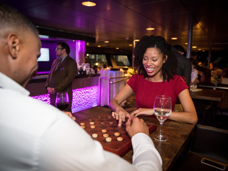 Paar spielt lächelnd Dame an der Bar der Dinner Cruise mit Weingläsern und lila Ambiente-Beleuchtung