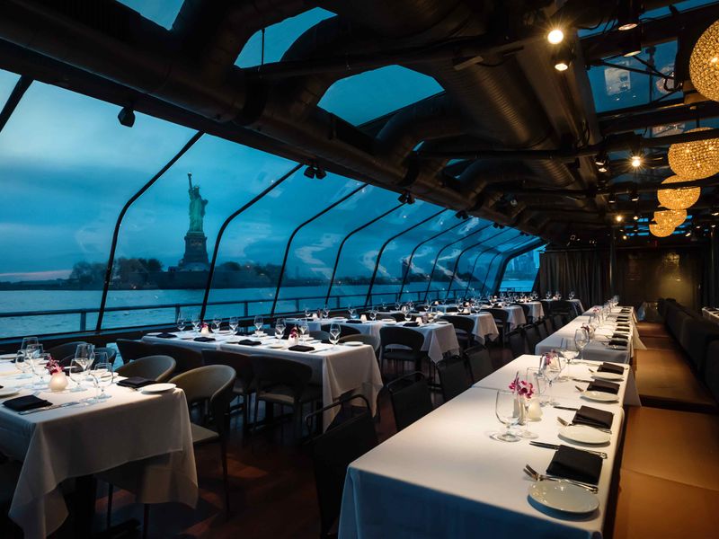 Elegant gedeckter Speisesaal auf einem Panoramaschiff in New York mit großen Glasfenstern und Blick auf die Freiheitsstatue.