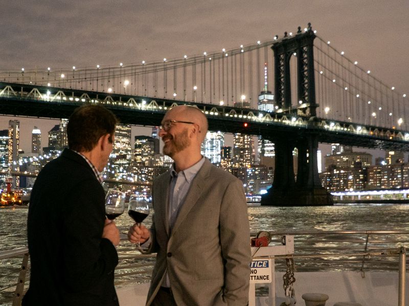 Zwei Männer mit Rotweingläsern unterhalten sich an Deck der Dinner Cruise vor der beleuchteten Manhattan Bridge bei Nacht