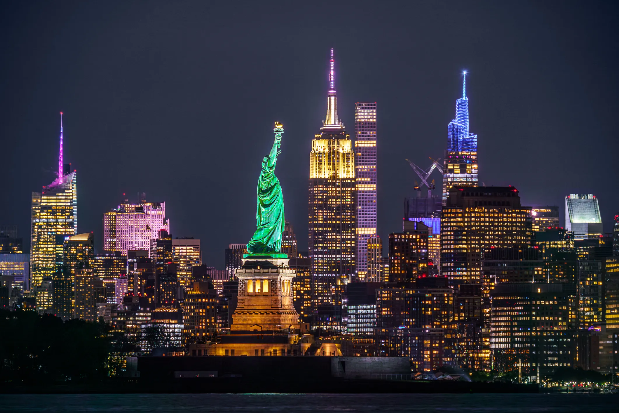 Die Freiheitsstatue in New York bei Nacht mit dem beleuchteten Empire State Building und der Manhattan-Skyline im Hintergrund