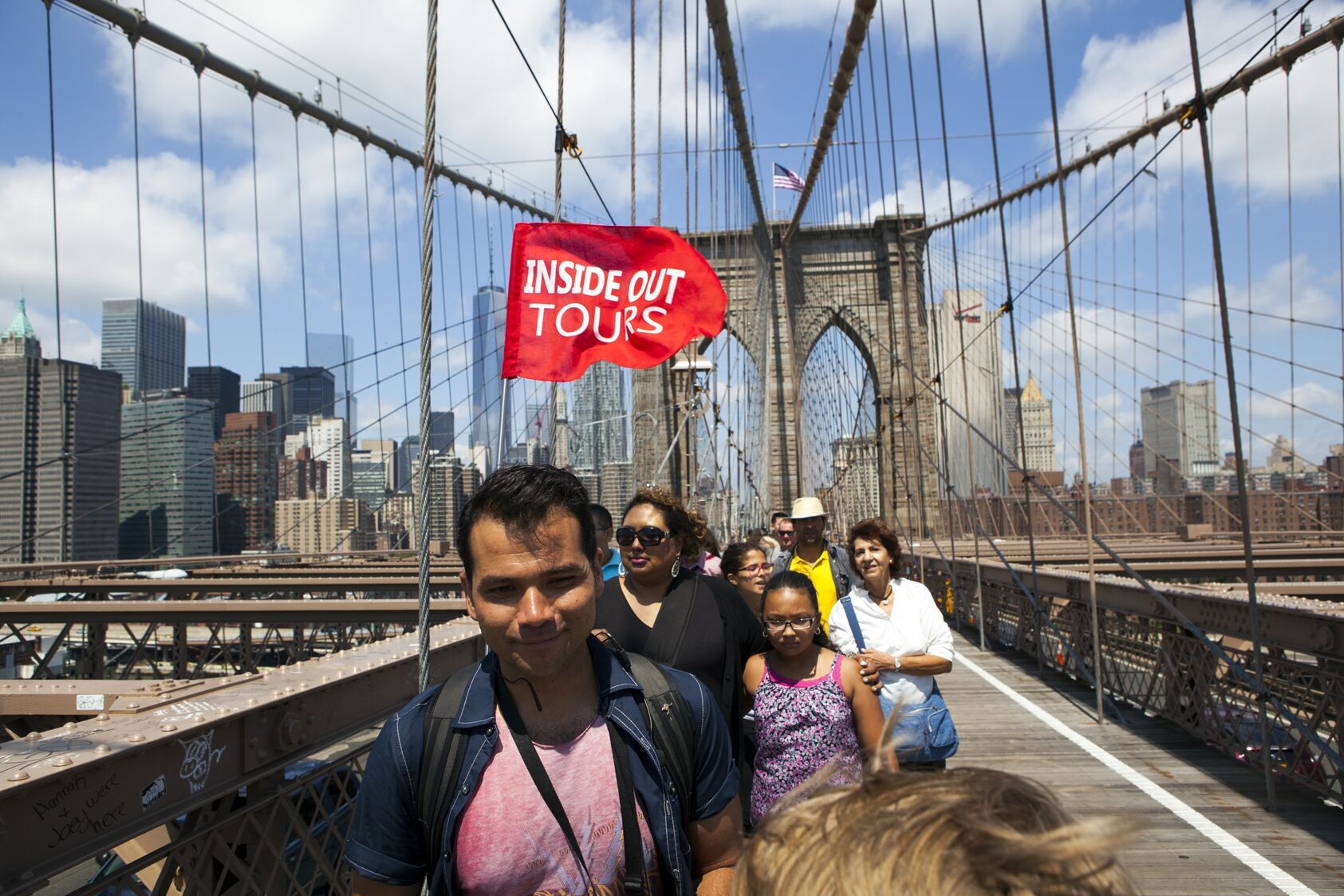 Eine Gruppe von Besuchern spaziert über den Fußgängerweg der Brooklyn Bridge, im Hintergrund sind die New Yorker Skyline und die Brückenpfeiler zu sehen.