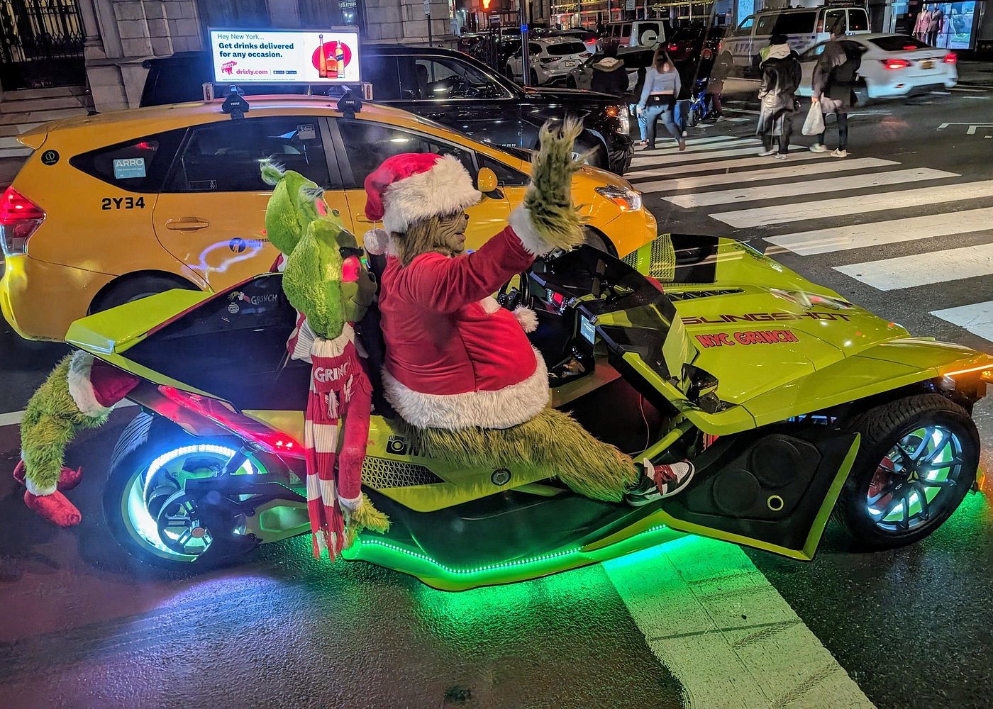 Als Grinch verkleidete Person mit Weihnachtsmütze in einem grün beleuchteten Fahrzeug neben einem gelben New Yorker Taxi.