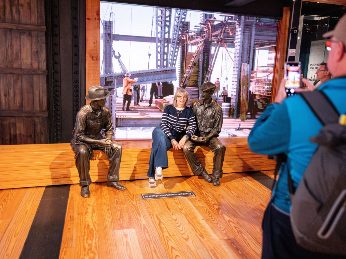 Eine Frau sitzt für ein Foto zwischen zwei historischen Bauarbeiter-Statuen in der interaktiven Ausstellung des Empire State Buildings.