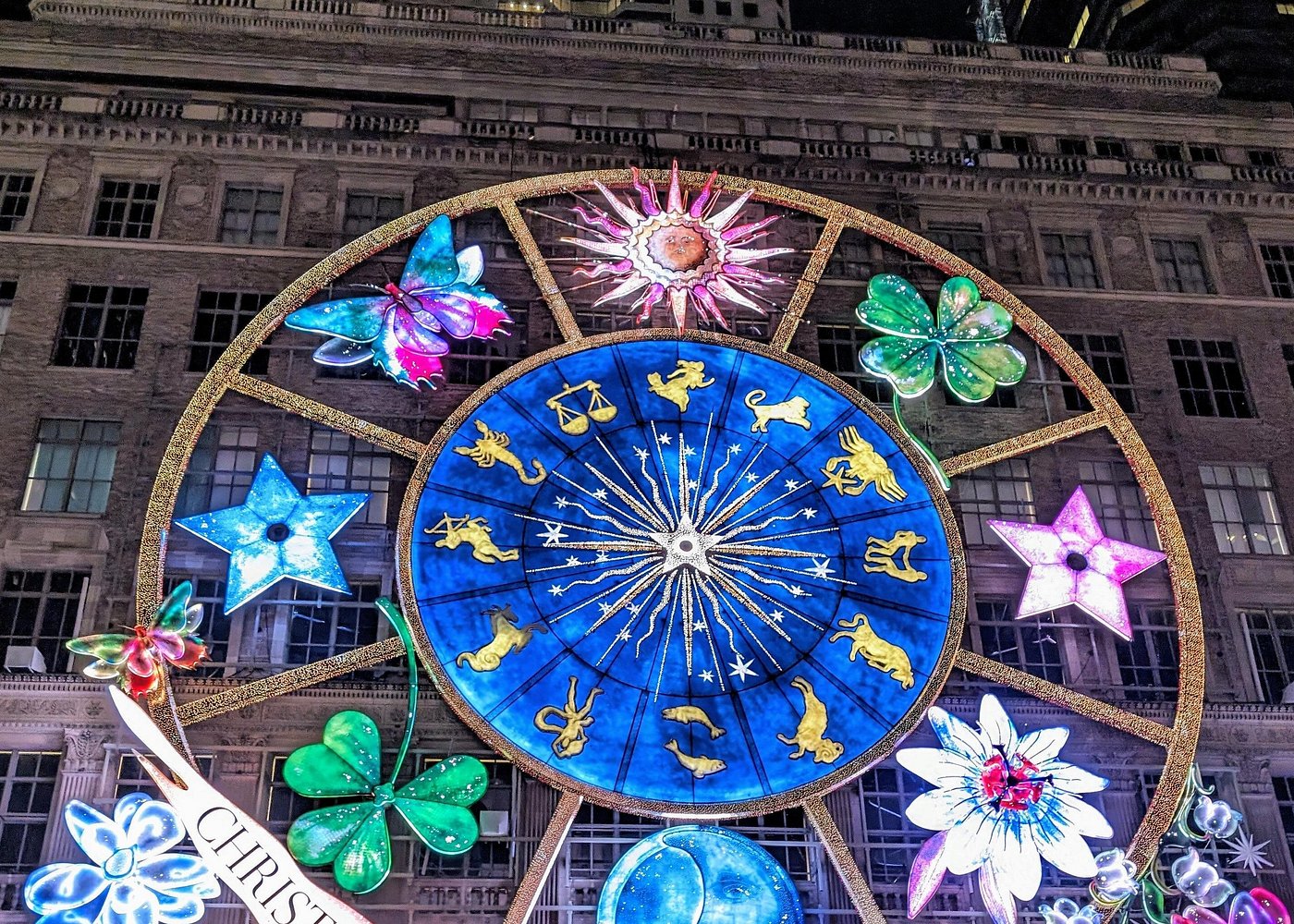 Farbenfrohe Weihnachtsbeleuchtung mit großem blauem Sternzeichenrad, Schmetterlingen und leuchtenden Blumen an einer Gebäudefassade.