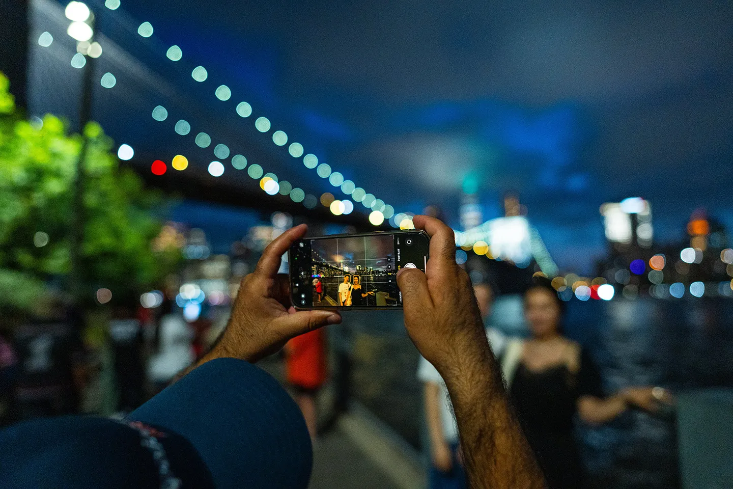 Hände halten ein Smartphone und fotografieren die beleuchtete Brooklyn Bridge und Manhattan-Skyline bei Nacht