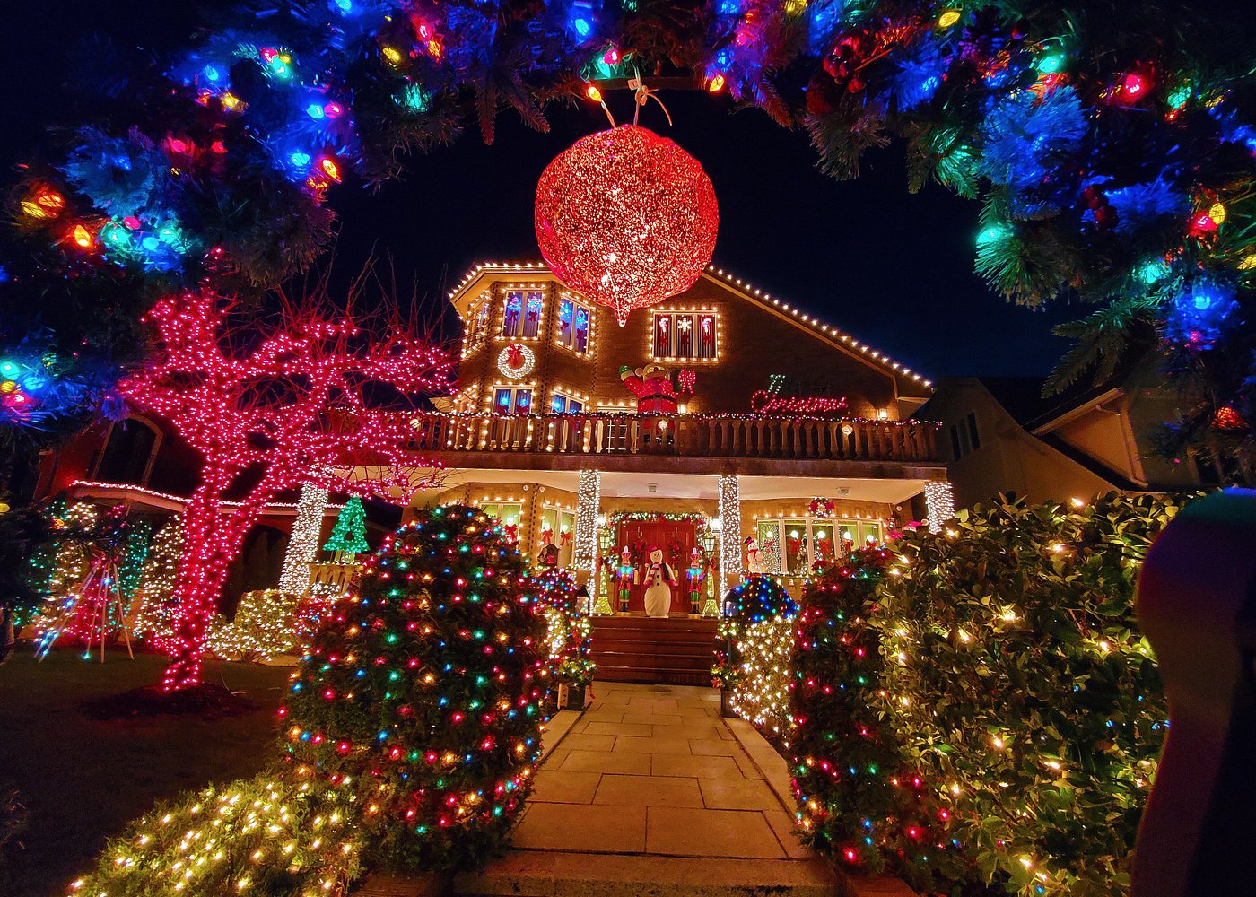 Weihnachtlich dekoriertes Haus in Dyker Heights mit leuchtenden Büschen, rotem Baum und großer Weihnachtskugel.
