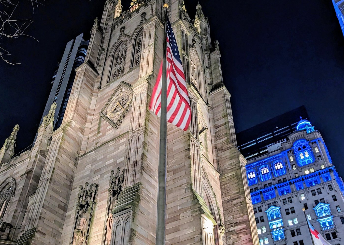 Beleuchtete neugotische Kirchenfassade in Manhattan mit amerikanischer Flagge und einem blau angestrahlten Gebäude im Hintergrund.