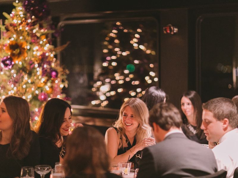 Gäste unterhalten sich beim festlichen Dinner an Bord eines Schiffes mit beleuchtetem Weihnachtsbaum im Hintergrund.