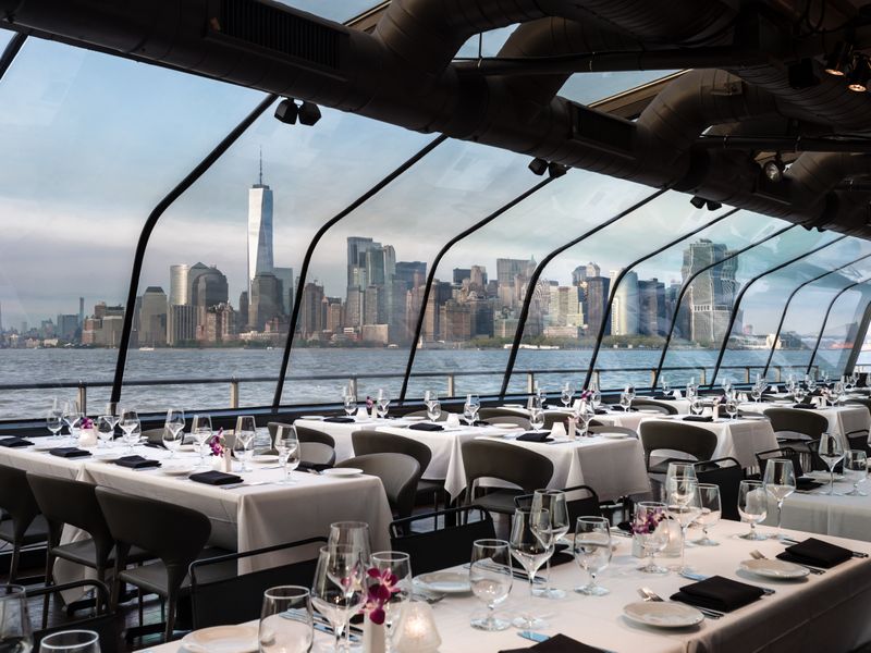 Blick durch den eleganten, verglasten Speisesaal eines Schiffes mit festlich gedeckten Tischen auf die Skyline mit dem One World Trade Center im Hintergrund.