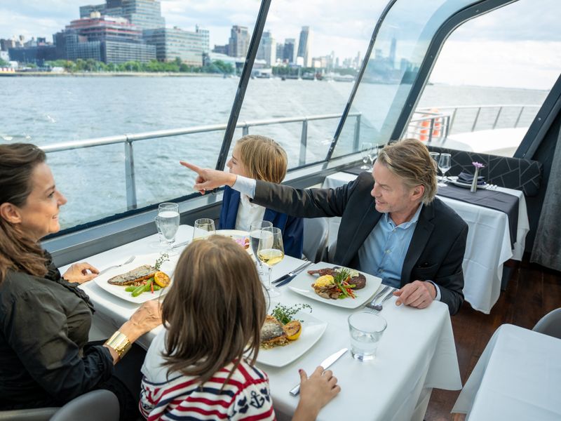 Eine Familie sitzt an einem gedeckten Tisch im verglasten Speisesaal eines Schiffs, während ein Mann aus dem Fenster auf die Skyline zeigt.