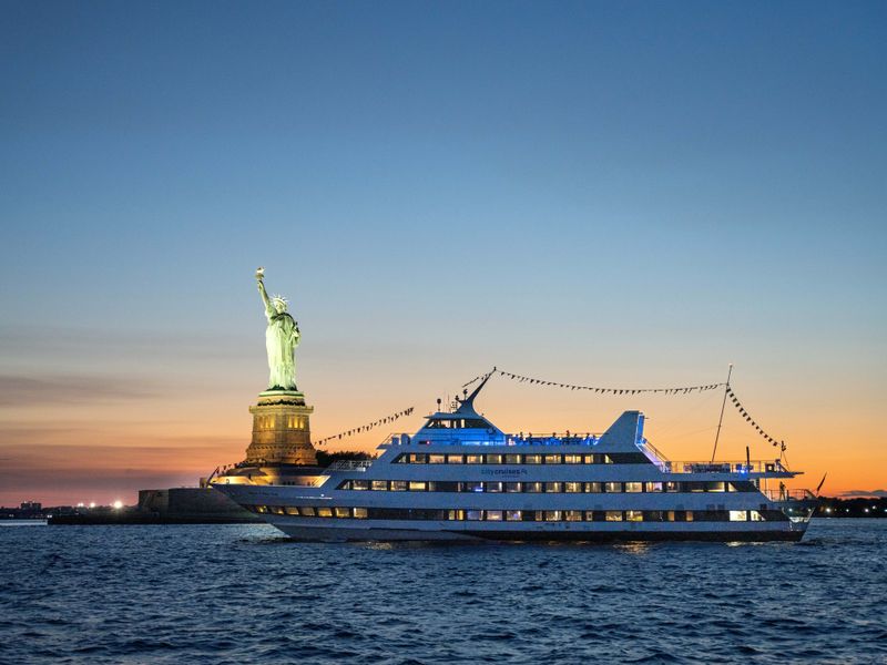 Großes Ausflugsschiff von City Cruises fährt bei Sonnenuntergang an der Freiheitsstatue im Hafen von New York vorbei.
