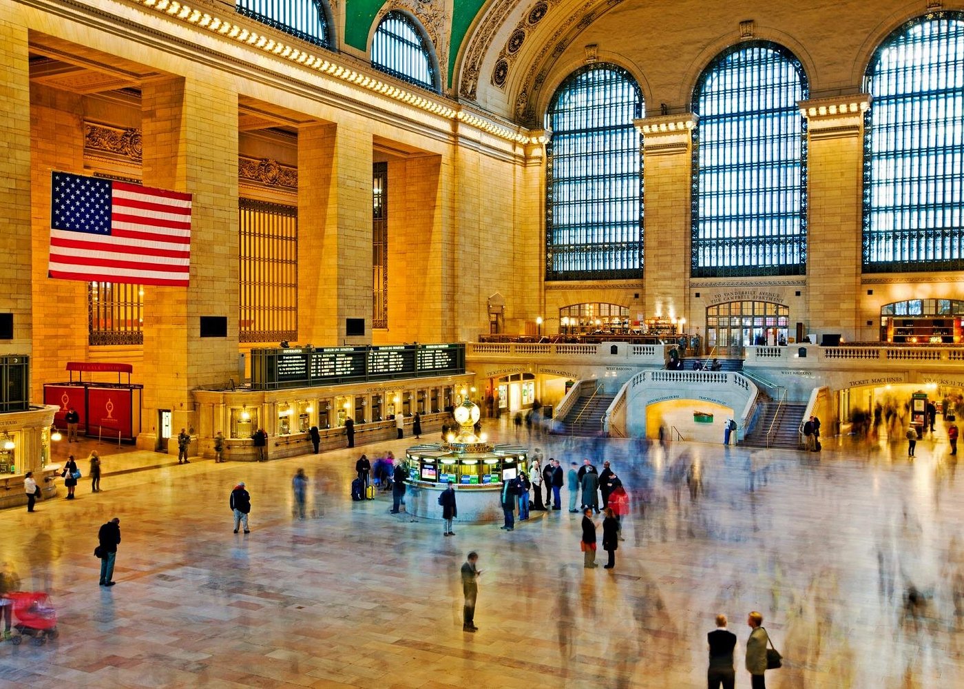Beeindruckende Haupthalle des Grand Central Terminal in Manhattan mit großen Bogenfenstern, amerikanischer Flagge und der berühmten Uhr.