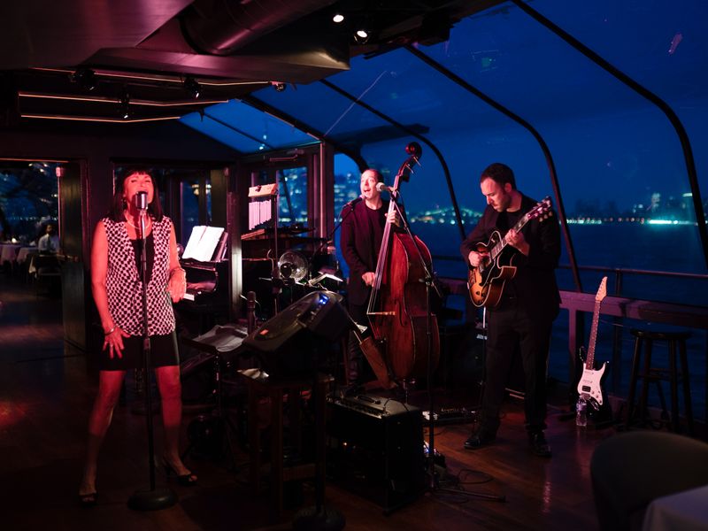 Jazzband mit Sängerin, Kontrabassist und Gitarrist spielt live auf einem Panoramaschiff mit Blick auf den New Yorker Hafen.