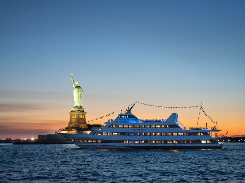 Beleuchtetes Dinner-Cruise-Schiff fährt bei Sonnenuntergang an der Freiheitsstatue vorbei