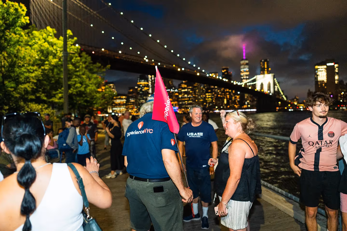 Reiseleiter mit Experience-NYC-Flagge spricht am Abend mit Teilnehmern einer Stadtführung vor der beleuchteten Brooklyn Bridge