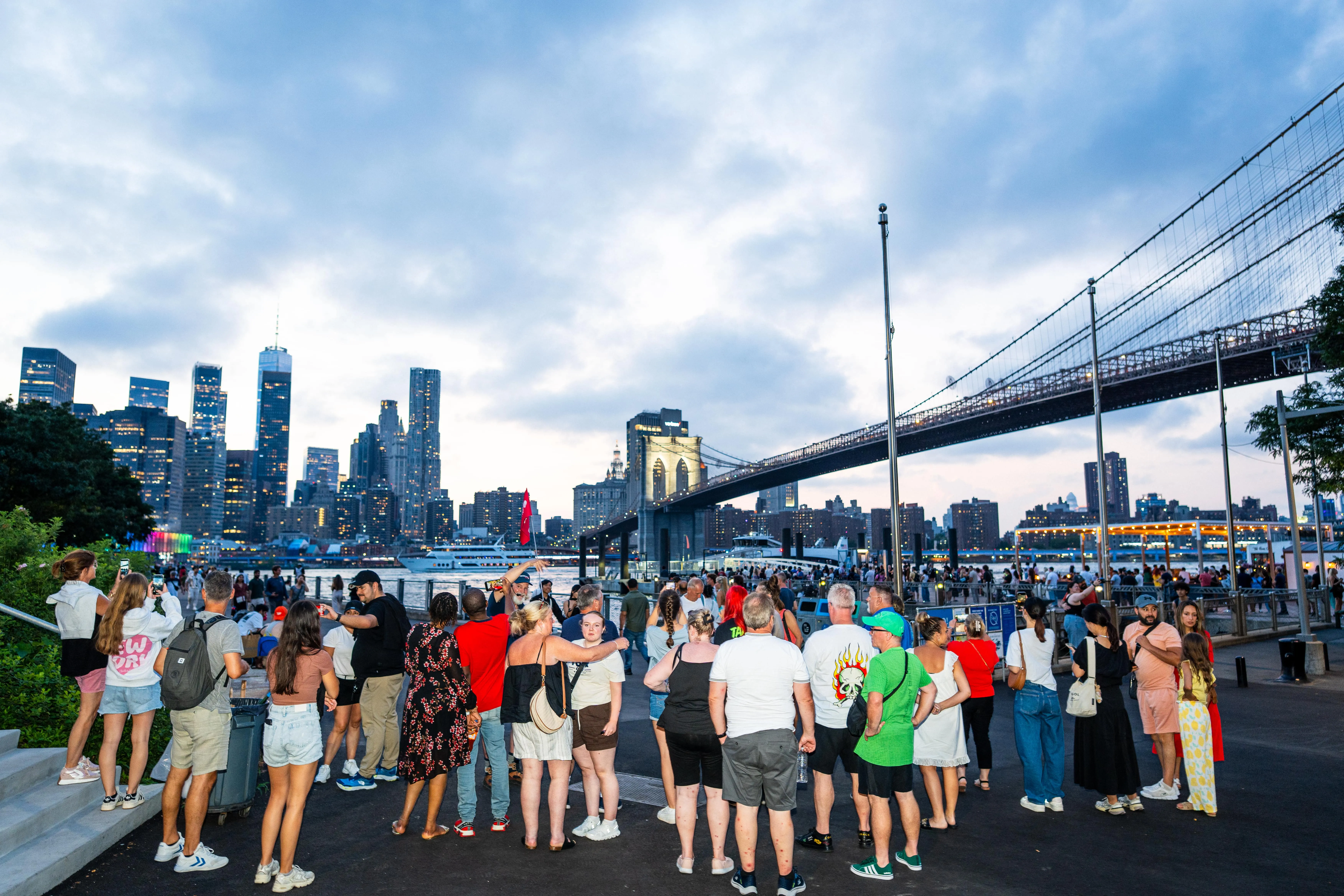 Große Touristengruppe steht auf der Uferpromenade und betrachtet die Brooklyn Bridge und die Skyline von Manhattan bei Dämmerung