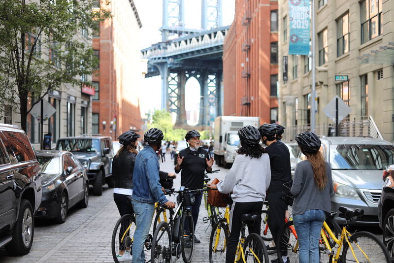 Gruppe bei einer geführten Fahrradtour durch DUMBO in Brooklyn mit der Manhattan Bridge im Hintergrund.