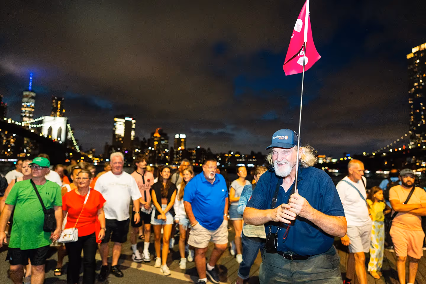Reiseleiter mit pinker Flagge und Experience-NYC-Cap führt eine Touristengruppe abends entlang des Brooklyn Bridge Park mit Skyline im Hintergrund