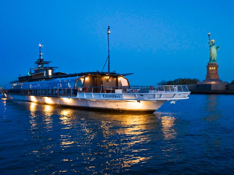 Beleuchtetes Panoramaschiff Celestial fährt in der Abenddämmerung an der Freiheitsstatue im Hafen von New York vorbei.