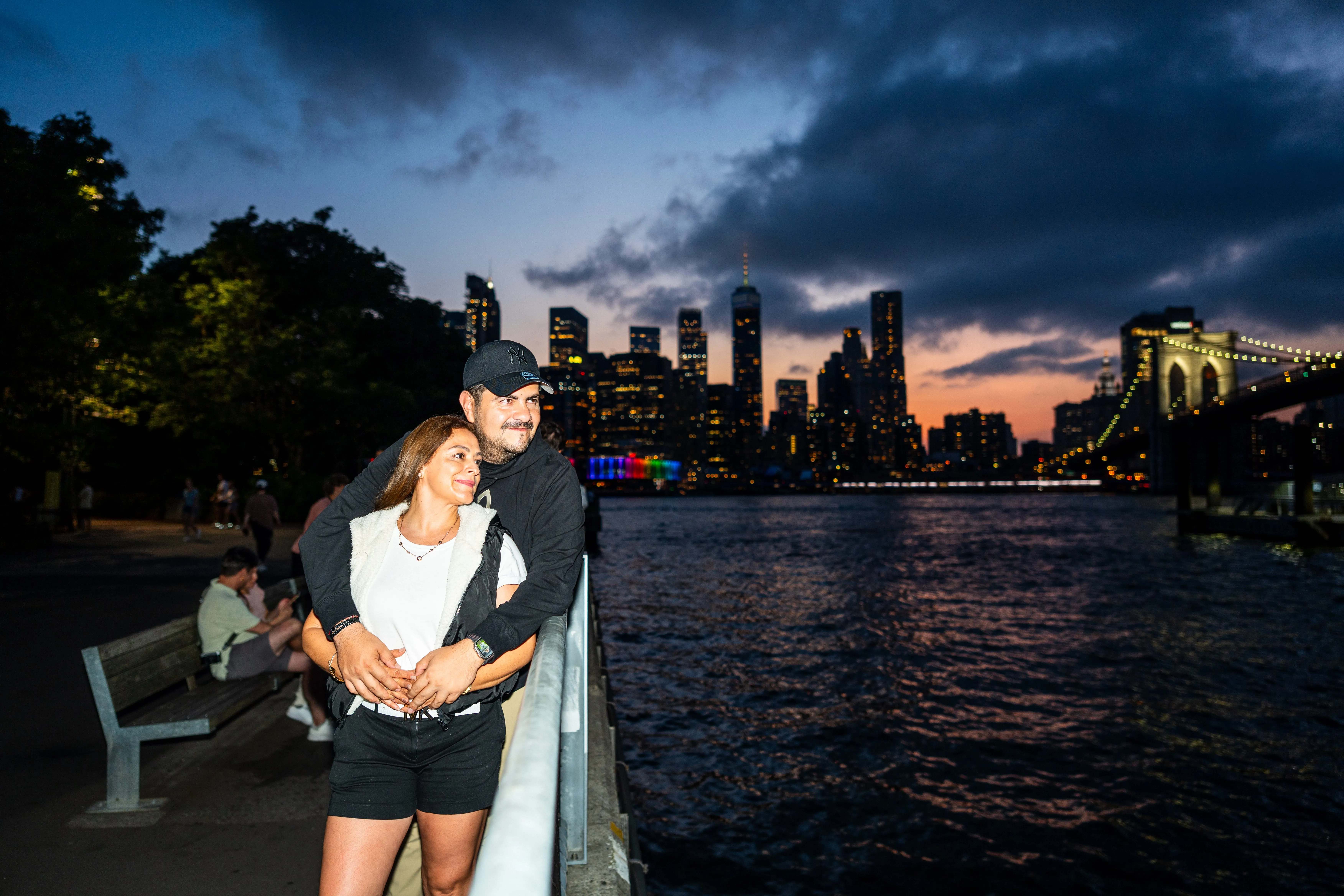 Paar umarmt sich am Geländer im Brooklyn Bridge Park und blickt auf die Manhattan-Skyline und Brooklyn Bridge bei Sonnenuntergang
