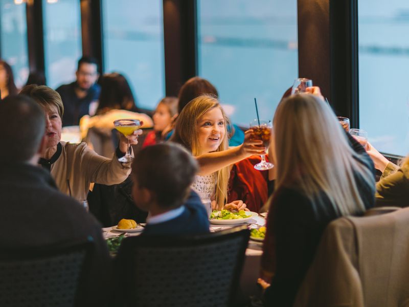 Familie stößt beim gemeinsamen Abendessen an Bord eines Panoramaschiffes mit Blick auf das Wasser an.