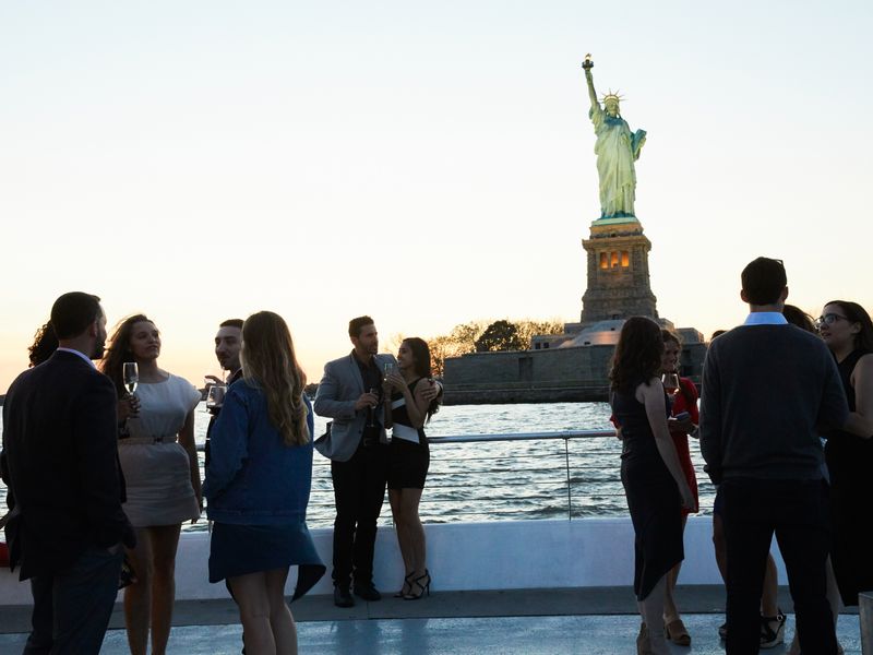Gäste genießen Getränke auf dem Deck eines Ausflugsschiffes mit direktem Blick auf die Freiheitsstatue bei Sonnenuntergang.