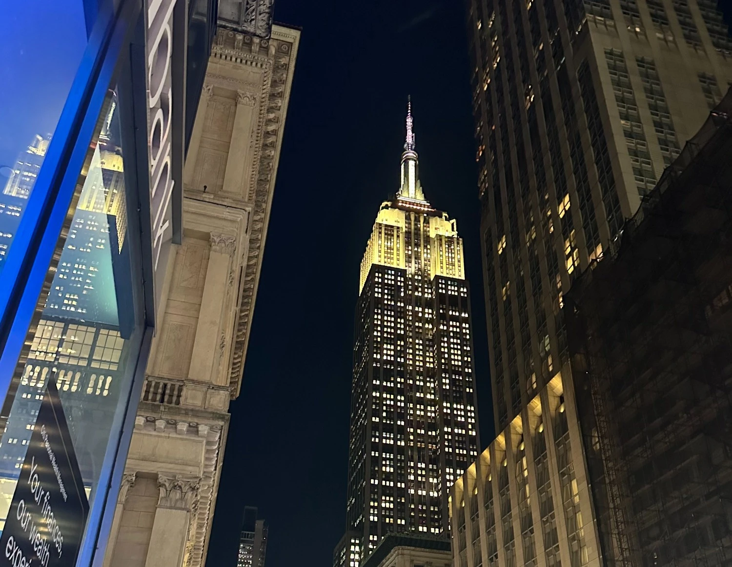 Blick von unten auf das beleuchtete Empire State Building bei Nacht, eingerahmt von umliegenden New Yorker Wolkenkratzern