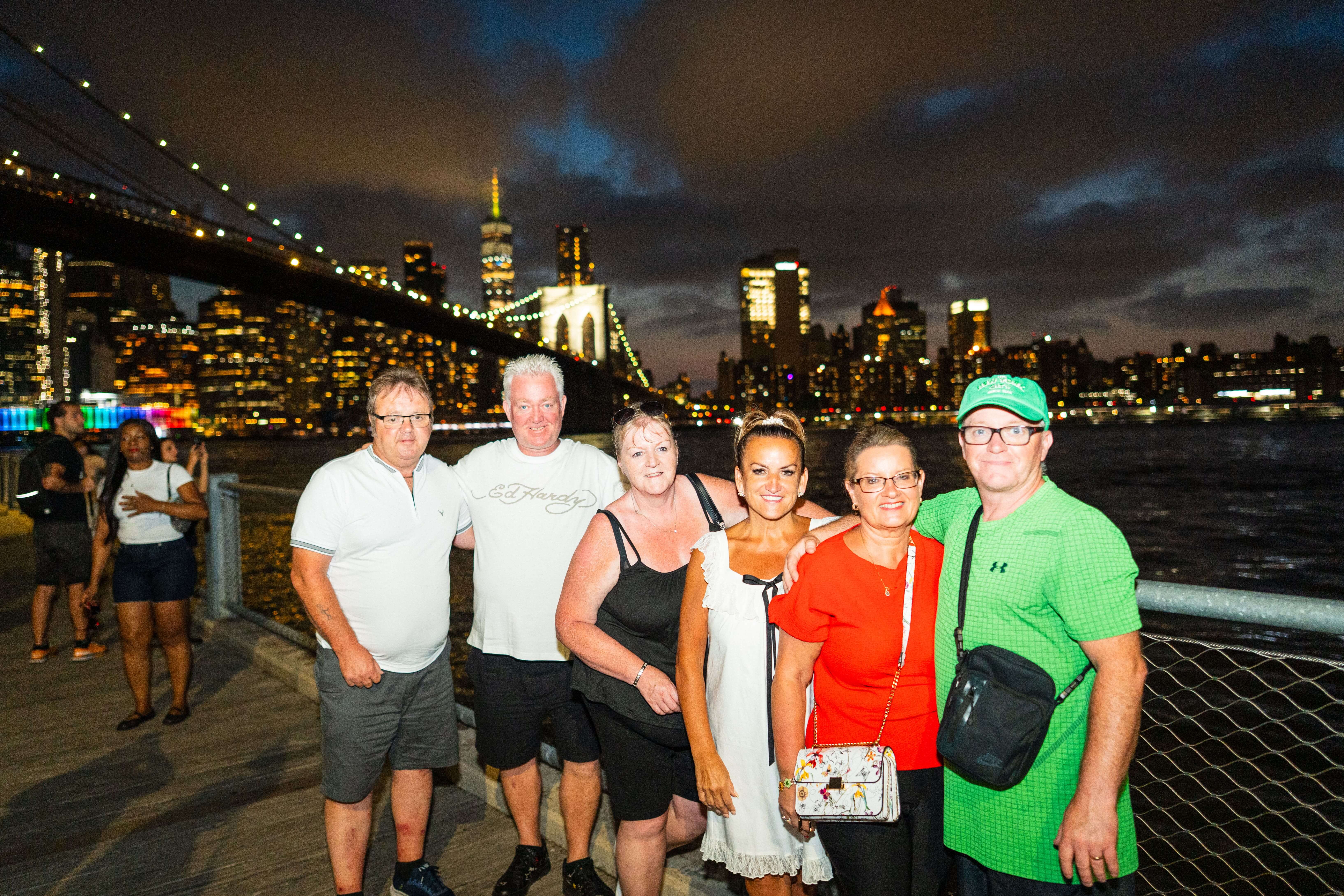 Sechs Touristen posieren lächelnd für ein Gruppenfoto auf der Brooklyn-Promenade mit Brooklyn Bridge und Manhattan-Skyline im Hintergrund