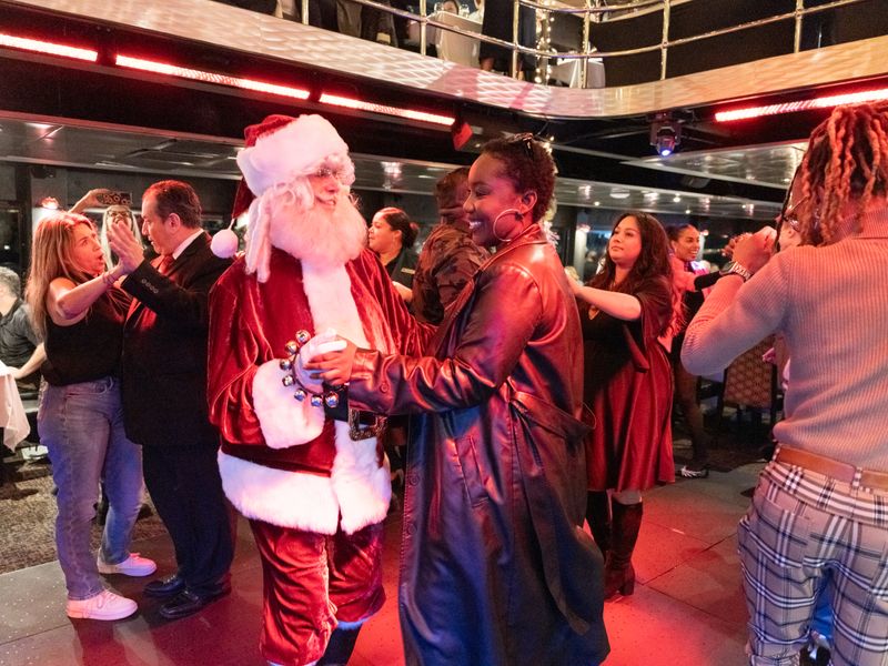 Weihnachtsmann tanzt mit Gästen auf einem festlich beleuchteten Ausflugsschiff während einer Weihnachtsfeier in New York.