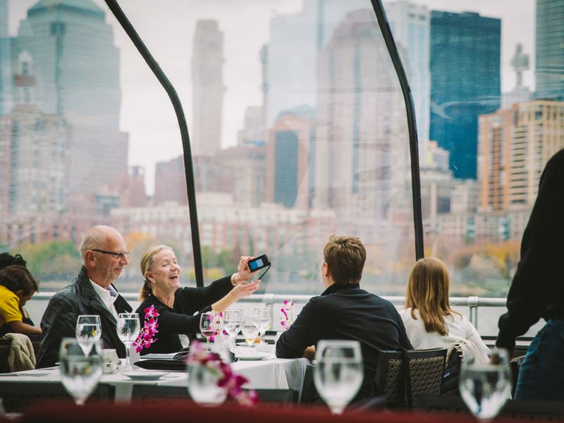 Gäste machen mit einer Kamera Fotos von der Skyline, während sie an einem Tisch unter dem Panorama-Glasdach eines Schiffs sitzen.