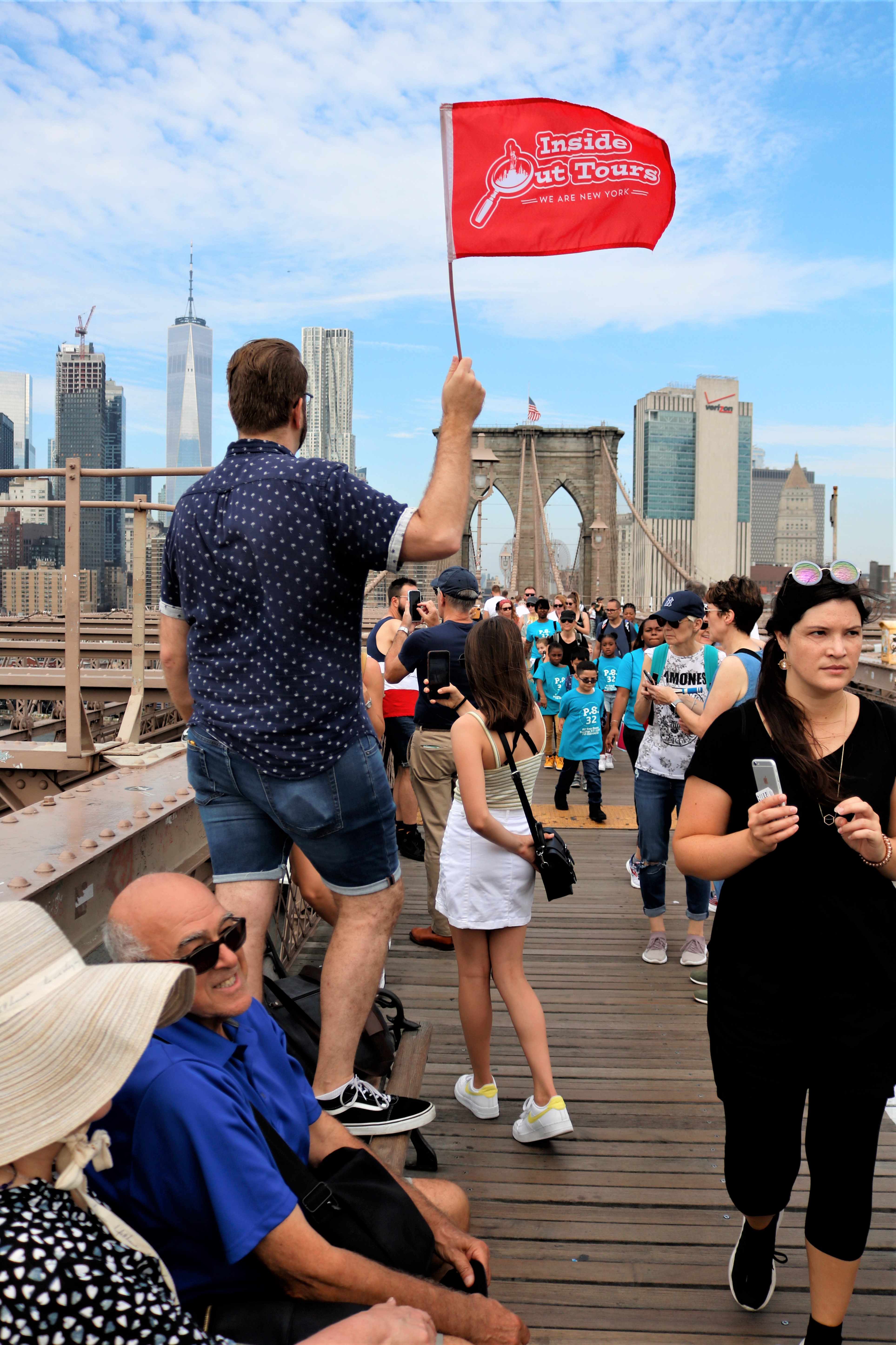 Eine Gruppe von Touristen geht über die Brooklyn Bridge, angeführt von einem Guide, der eine rote Fahne mit der Aufschrift „Inside Out Tours“ in die Höhe hält.