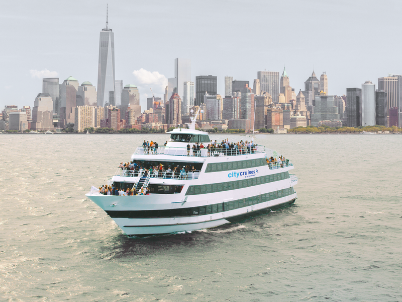 Großes Ausflugsschiff von City Cruises fährt über den New Yorker Hafen mit der Skyline von Lower Manhattan und dem One World Trade Center im Hintergrund.